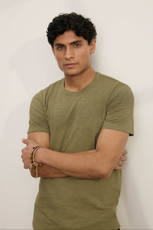 A man with short curly hair stands against a plain light-colored wall, arms crossed, wearing the AMARO TEE by Velvet by Graham & Spencer—an olive green Peruvian cotton crew neck t-shirt.