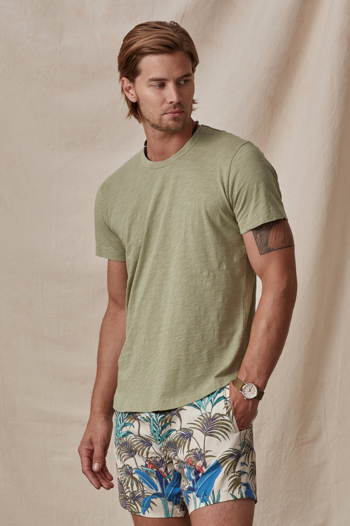 A man in a green Velvet by Graham & Spencer AMARO TEE and tropical print shorts standing against a beige backdrop. He is also wearing a watch.-36752671539393