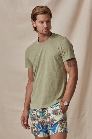 A man in a green Velvet by Graham & Spencer AMARO TEE and tropical print shorts standing against a beige backdrop. He is also wearing a watch.
