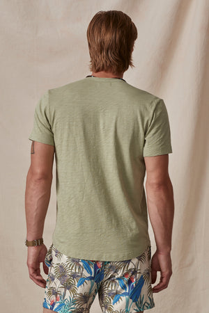 A man viewed from behind wearing a green AMARO TEE by Velvet by Graham & Spencer with a curved hemline and colorful tropical print shorts.