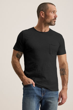 A bearded man wears the CHAD TEE from Velvet by Graham & Spencer, a black T-shirt with raw edge details and a classic crew neckline. He pairs it with blue jeans, standing against a plain backdrop while gazing thoughtfully to the side.