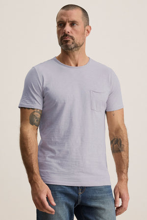 A man with short hair and tattoos wears the CHAD TEE from Velvet by Graham & Spencer, a light gray, soft Peruvian cotton T-shirt featuring a crew neckline and chest pocket. Paired with blue jeans, he gazes to the side against a neutral background.