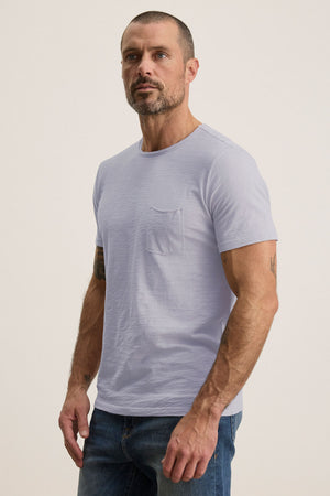 A man with a beard wears the CHAD TEE from Velvet by Graham & Spencer—a light gray, crew neck T-shirt with a chest pocket, crafted from soft Peruvian cotton—and stands against a plain background in blue jeans.
