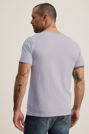 A tattooed man stands facing away against a plain background, wearing the CHAD TEE in light gray from Velvet by Graham & Spencer, made of soft Peruvian cotton with a crew neckline, paired with jeans.