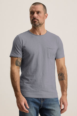 A man with short hair and a beard wears the light gray CHAD TEE by Velvet by Graham & Spencer with a crew neckline and blue jeans, standing against a plain background.