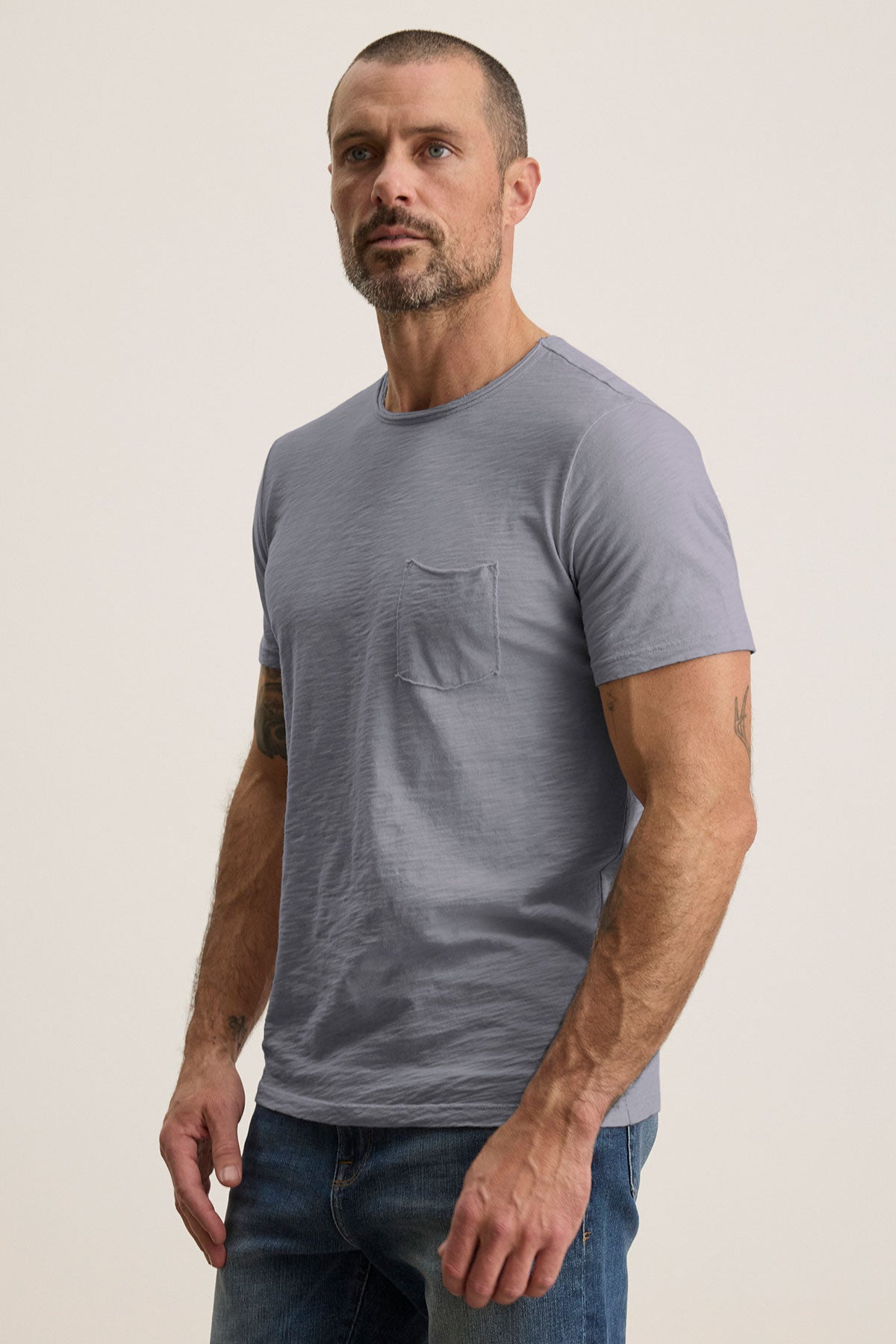 A man with short hair and a beard wears the CHAD TEE by Velvet by Graham & Spencer—a light grey Peruvian cotton t-shirt with a chest pocket and crew neck—paired with blue jeans, standing against a plain background.
