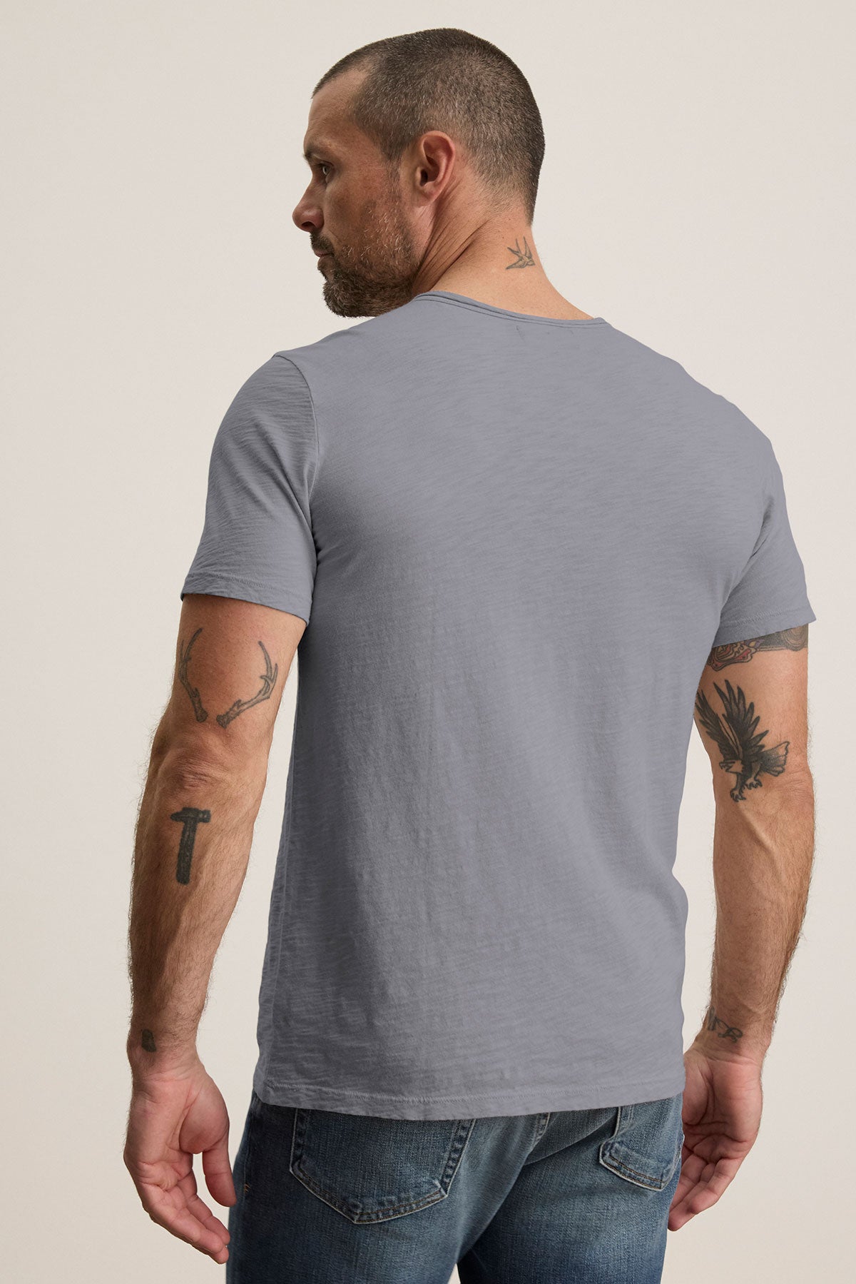 A man with short hair and arm tattoos wears the light gray CHAD TEE by Velvet by Graham & Spencer, featuring a chest patch pocket, paired with blue jeans, standing with his back to the camera.