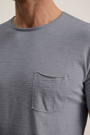 A person is shown from neck to upper torso wearing the CHAD TEE by Velvet by Graham & Spencer, a light gray short-sleeve crewneck t-shirt with a raw-edged chest patch pocket.