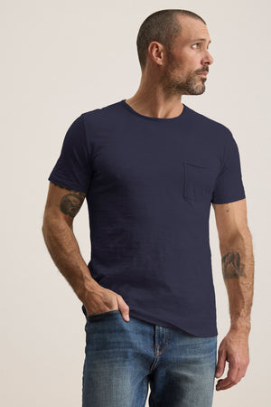 A tattooed, bearded man wears the Velvet by Graham & Spencer CHAD TEE in navy, featuring a classic crew neckline and raw edge details, paired with jeans, as he looks to his right.