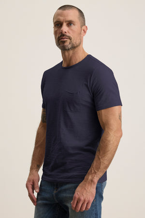 A bearded man with short hair stands against a plain background, wearing the Velvet by Graham & Spencer CHAD TEE—dark blue Peruvian cotton with a crew neckline—and jeans.
