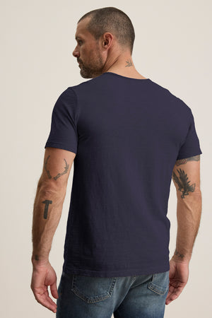 Man with short hair and tattoos on arms and neck wearing the Velvet by Graham & Spencer CHAD TEE in navy blue, featuring a crew neckline, paired with jeans, viewed from behind.