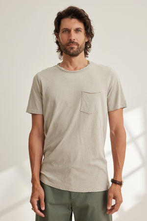 A man with medium-length brown hair and a beard wears the CHAD TEE by Velvet by Graham & Spencer—a light beige Peruvian cotton crew-neck short-sleeve shirt—paired with green pants, standing indoors against a plain background.