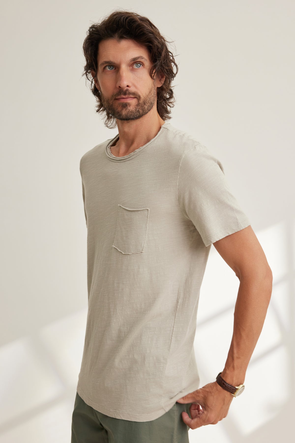 A man with wavy hair and a beard wears the CHAD TEE by Velvet by Graham & Spencer—a light beige Peruvian cotton short-sleeve t-shirt with a chest patch pocket and crew neckline—standing indoors against a neutral background.-39096572936385