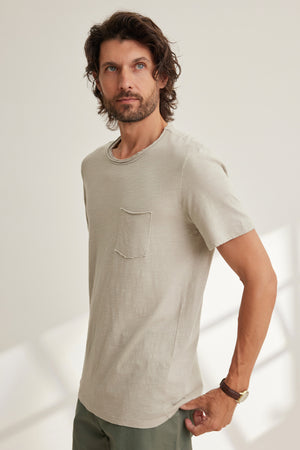 A man with wavy hair and a beard wears the CHAD TEE by Velvet by Graham & Spencer—a light beige Peruvian cotton short-sleeve t-shirt with a chest patch pocket and crew neckline—standing indoors against a neutral background.