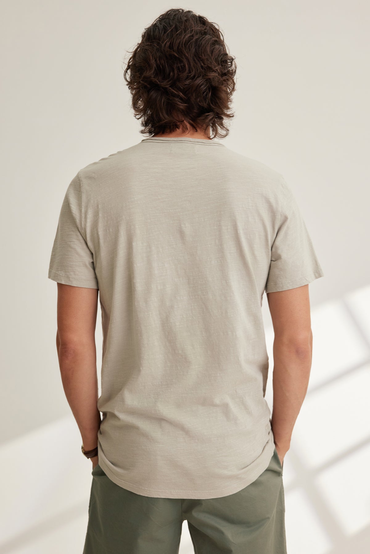 A person with medium-length wavy hair stands indoors, facing away, wearing the CHAD TEE by Velvet by Graham & Spencer in light beige with a crew neckline and olive green pants.-39096572969153