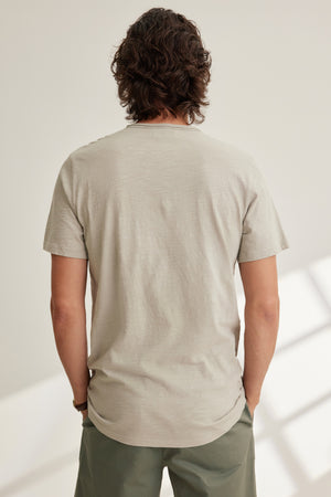 A person with medium-length wavy hair stands indoors, facing away, wearing the CHAD TEE by Velvet by Graham & Spencer in light beige with a crew neckline and olive green pants.