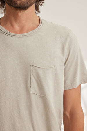 Man wearing the CHAD TEE by Velvet by Graham & Spencer, a light beige short-sleeve crewneck t-shirt with a raw-edge chest patch pocket, shown from shoulders to mid-torso.
