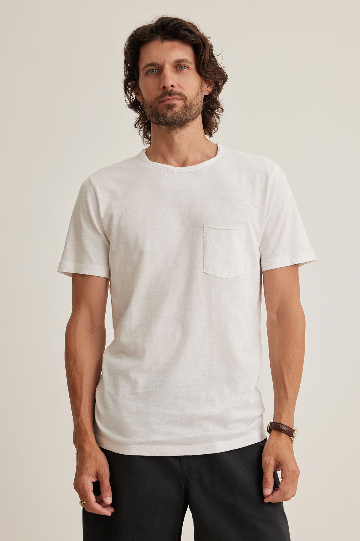 A man with long hair wearing a CHAD TEE from Velvet by Graham & Spencer crafted from Peruvian cotton, black pants, and a wristwatch stands against a plain background. The white t-shirt features raw edge details and a classic crew neckline for an effortlessly stylish look.