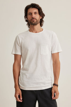 A man with long hair wearing a CHAD TEE from Velvet by Graham & Spencer crafted from Peruvian cotton, black pants, and a wristwatch stands against a plain background. The white t-shirt features raw edge details and a classic crew neckline for an effortlessly stylish look.