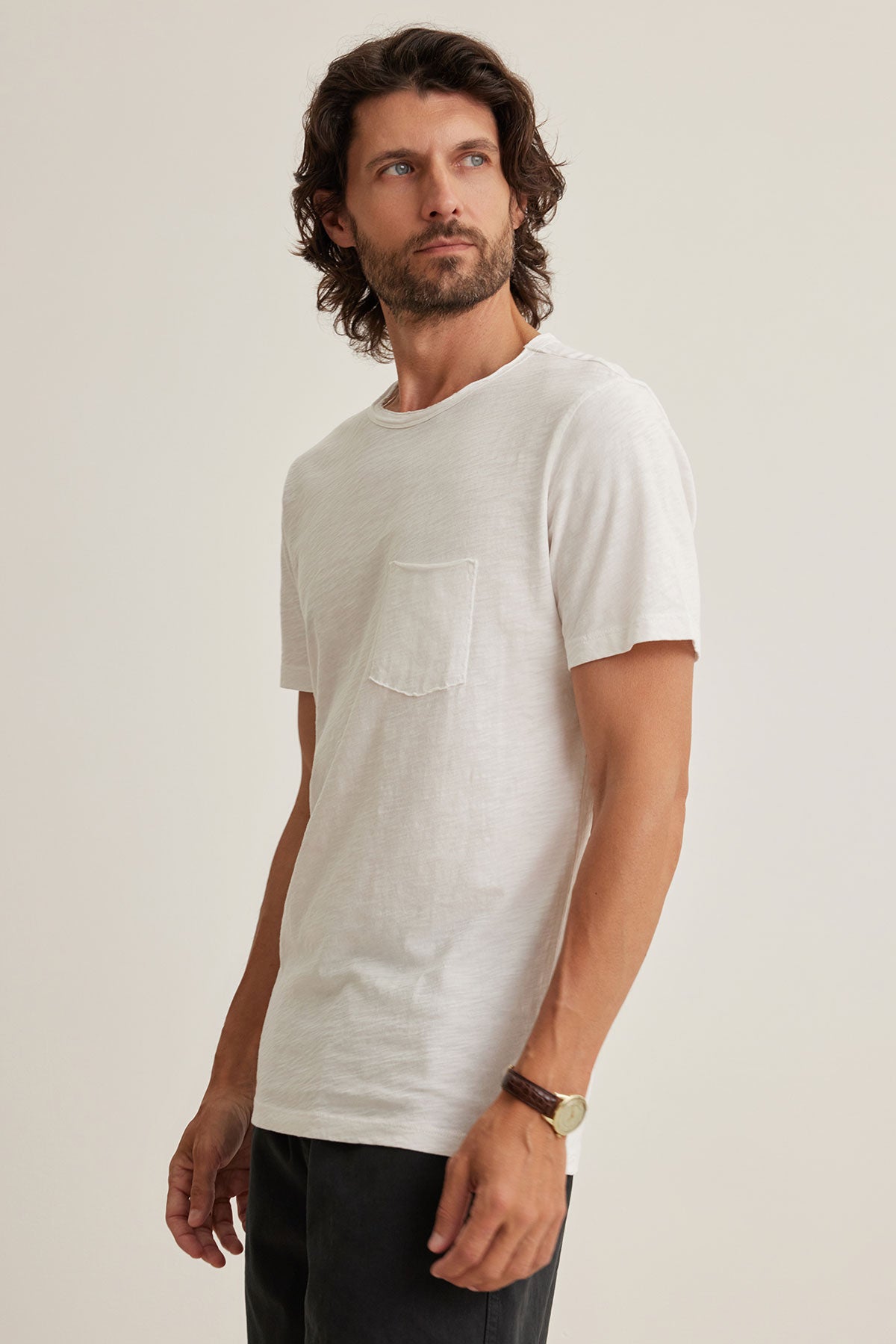 A long-haired man in a soft Peruvian cotton CHAD TEE by Velvet by Graham & Spencer, paired with black pants showcasing raw edge details, gazes sideways against a plain backdrop.-38877622304961