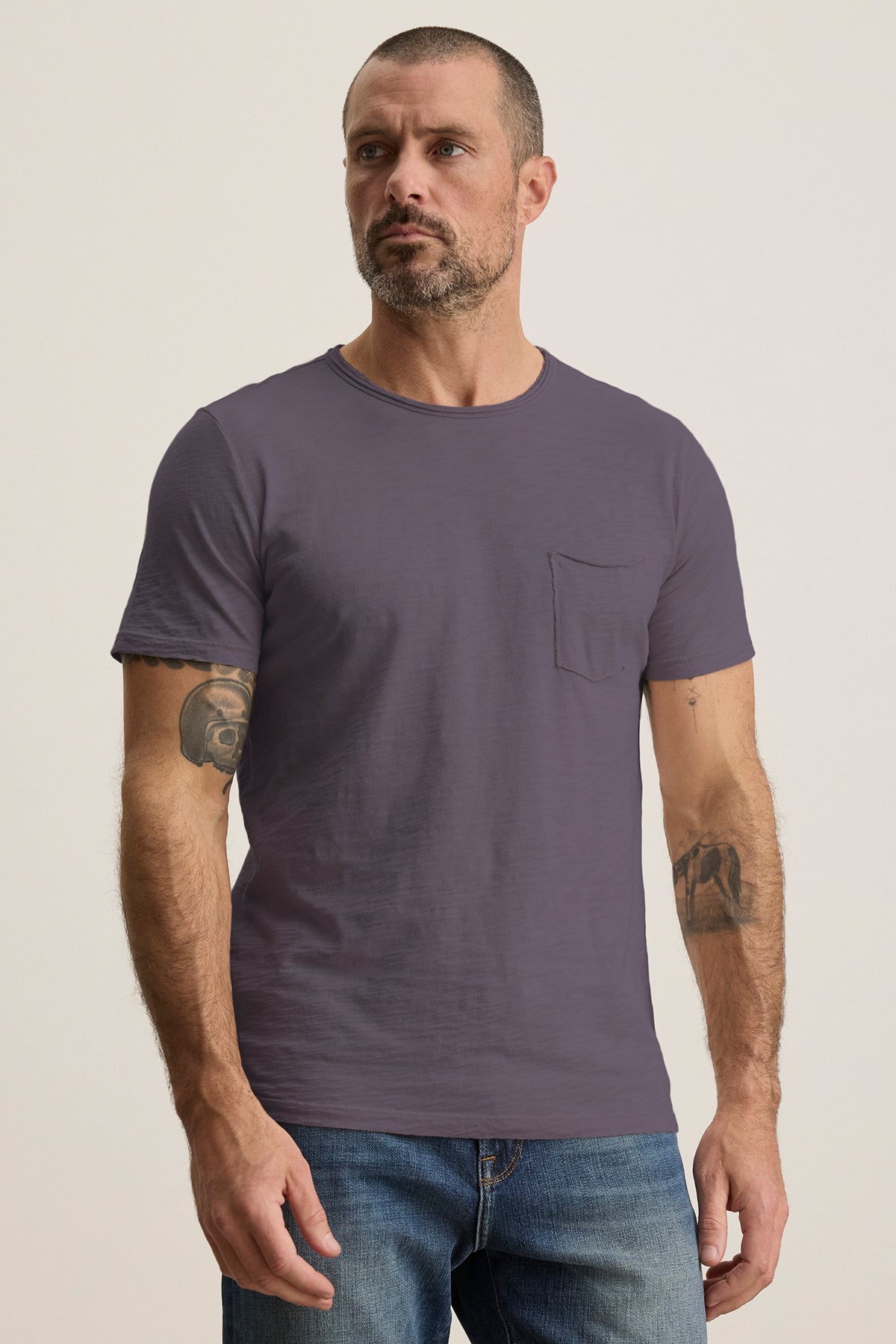 A tattooed man stands against a neutral background wearing the CHAD TEE, a plain purple crew-neck T-shirt by Velvet by Graham & Spencer, paired with blue jeans.-38807474929857