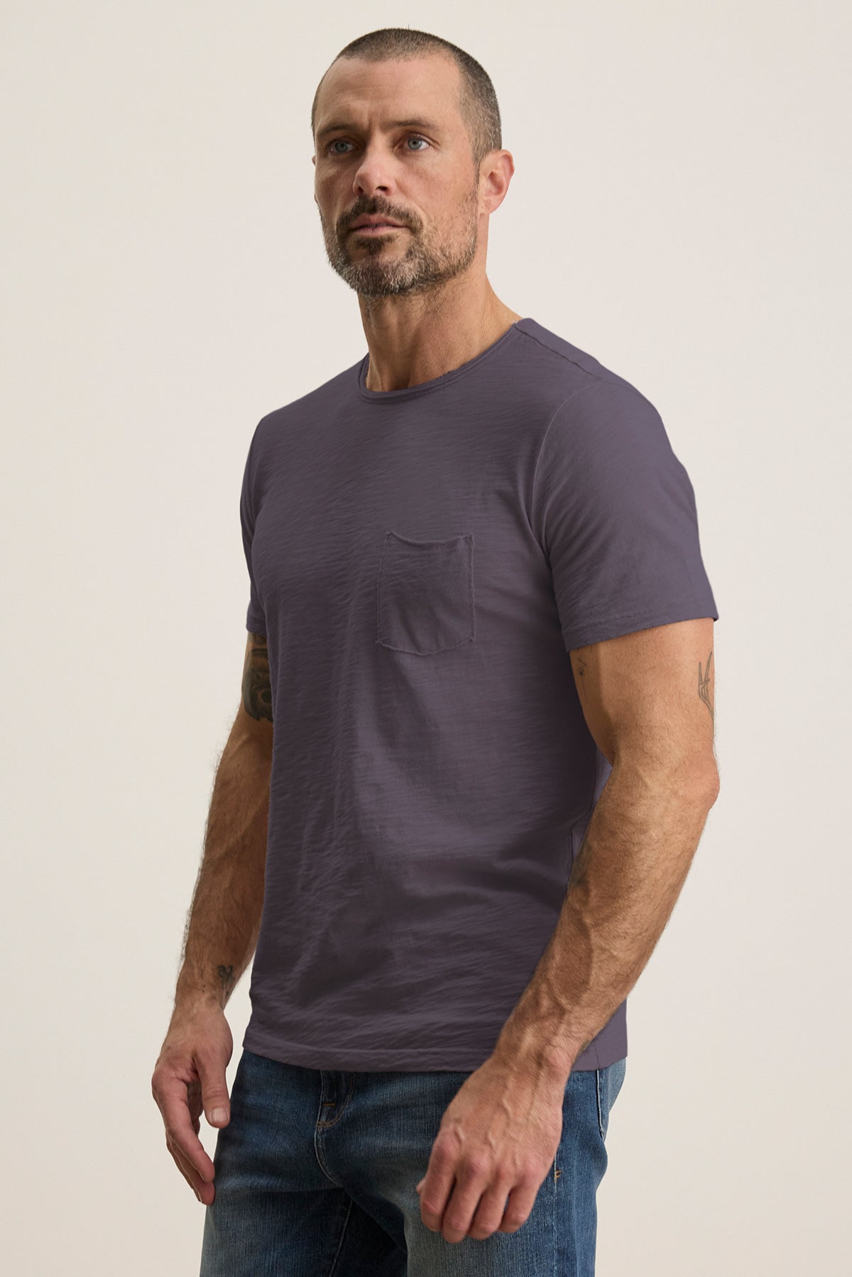 A person with short hair and a beard wears Velvet by Graham & Spencer's CHAD TEE in gray, made from soft Peruvian cotton, with a crew neckline and chest patch pocket, paired with jeans against a neutral background.-38807474962625