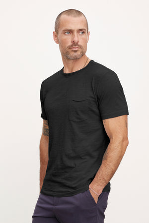 A man with a short beard in a Velvet by Graham & Spencer CHAD TEE with raw-edge details and purple pants, standing against a plain background.