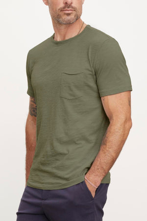 A man with a cropped haircut and beard, wearing the CHAD TEE by Velvet by Graham & Spencer with a classic crew neckline and dark pants, stands looking off to the side. His left arm has visible tattoos, while the textured cotton slub of his shirt adds a unique touch.