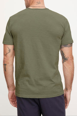Man with a shaved head and tattoos on his neck and arms, sporting an olive green short-sleeve CHAD TEE made from textured cotton slub by Velvet by Graham & Spencer, and dark pants, shown from the back.