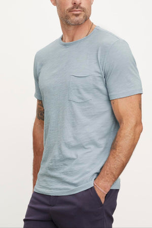 A person with short hair and a beard wears a light blue CHAD TEE by Velvet by Graham & Spencer with raw edge details and dark pants, looking to the side. The classic crew neckline t-shirt has a chest pocket made of textured cotton slub. The background is plain and light-colored.