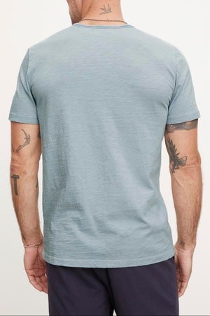 A man with short hair and tattoos on both arms is shown from the back, wearing a light blue CHAD TEE by Velvet by Graham & Spencer made of textured cotton slub with raw edge details, paired with dark pants.
