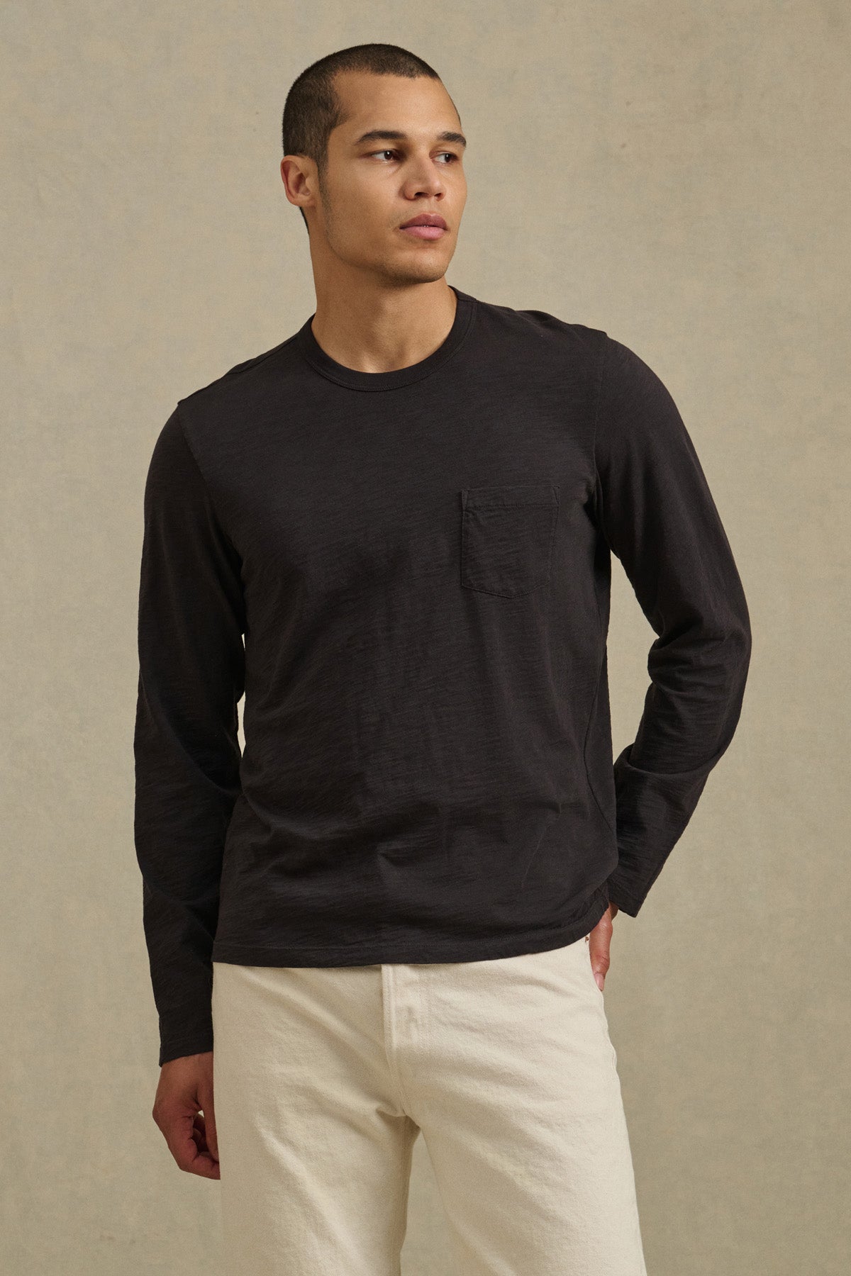 A man wears the DALTON TEE by Velvet by Graham & Spencer—a black cotton slub long-sleeve tee with a chest pocket—paired with off-white pants, standing against a neutral background for effortless everyday style.-40850714755265