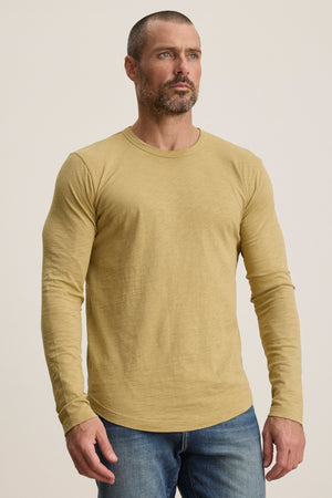 A man with short hair and a beard wears the KAI TEE by Velvet by Graham & Spencer—a light yellow Peruvian cotton long sleeve crewneck tee—paired with blue jeans, standing against a plain light background.