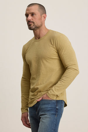 A man with short hair and a beard wears the KAI TEE by Velvet by Graham & Spencer, a mustard yellow Peruvian cotton long sleeve crewneck, paired with blue jeans, standing against a plain light background.