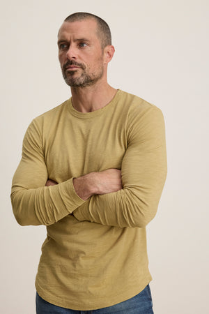 A man with short hair and a beard wears the Velvet by Graham & Spencer KAI TEE, a light brown Peruvian cotton long sleeve crewneck tee, paired with blue jeans, standing with arms crossed against a plain background.