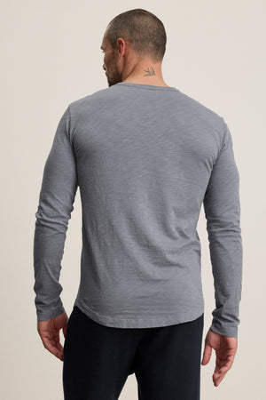 A man with short hair and a neck tattoo, wearing the Velvet by Graham & Spencer KAI TEE—a gray long sleeve crewneck—stands facing away from the camera against a plain background.