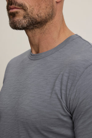 A close-up of a man wearing the light gray KAI TEE by Velvet by Graham & Spencer, showing his neck, lower face, and part of his shoulder against a plain background.
