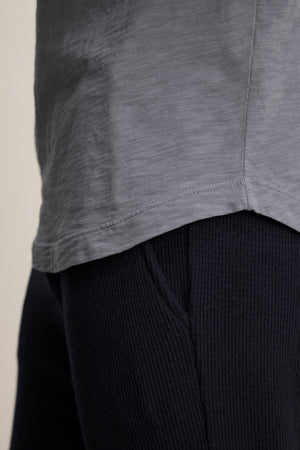 Close-up of a person wearing the Velvet by Graham & Spencer KAI TEE in light gray and black textured pants, focusing on the shirt's lower section and the waistband area of the pants.