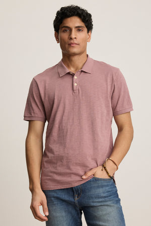 A man faces forward in the Velvet by Graham & Spencer NIKO POLO, featuring mauve Peruvian cotton short sleeves and blue jeans, with one hand in his pocket against a plain background.