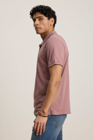 A man stands sideways against a plain light background, showcasing the effortless versatility of the Velvet by Graham & Spencer NIKO POLO in mauve paired with blue jeans.