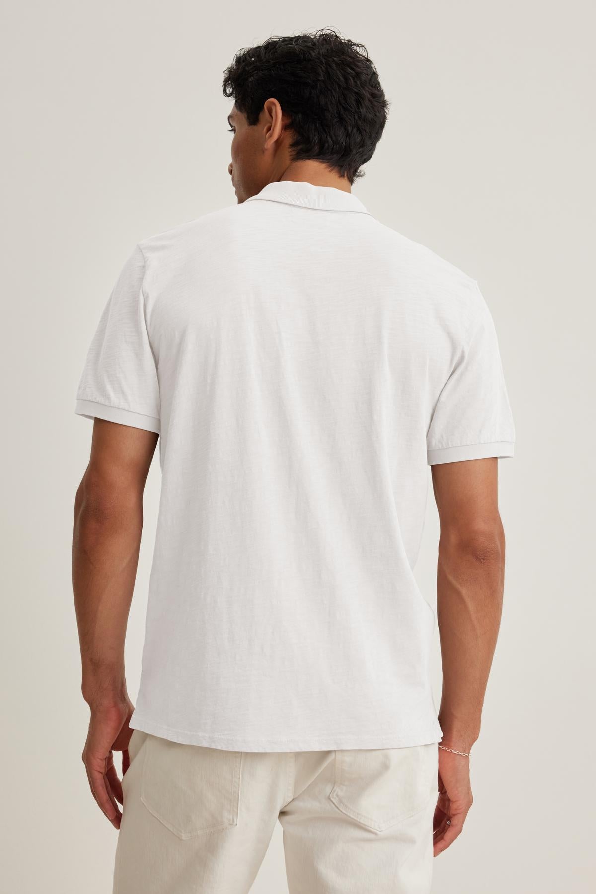 A man with short hair stands facing away, wearing a NIKO POLO by Velvet by Graham & Spencer made from soft Peruvian cotton and light-colored pants, epitomizing versatile wear against a neutral background.-38658660499649