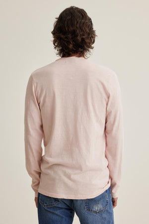 A person with curly hair stands facing away, wearing a Velvet by Graham & Spencer SIMEON TEE in light pink made from soft Peruvian cotton and blue jeans with raw-edge details against a plain background.