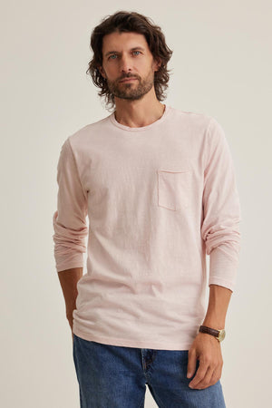 A bearded individual in the Velvet by Graham & Spencer SIMEON TEE, a light pink Peruvian cotton long-sleeve shirt with raw edges, paired with blue jeans, stands against a plain backdrop, hands in pockets, embodying California heritage style.