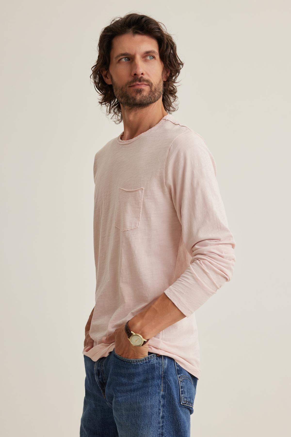 A man with curly hair stands with hands in pockets, wearing a Velvet by Graham & Spencer SIMEON TEE in light pink, featuring raw-edge details, alongside jeans. He looks to the side.-38643350208705
