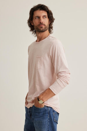 A man with curly hair stands with hands in pockets, wearing a Velvet by Graham & Spencer SIMEON TEE in light pink, featuring raw-edge details, alongside jeans. He looks to the side.