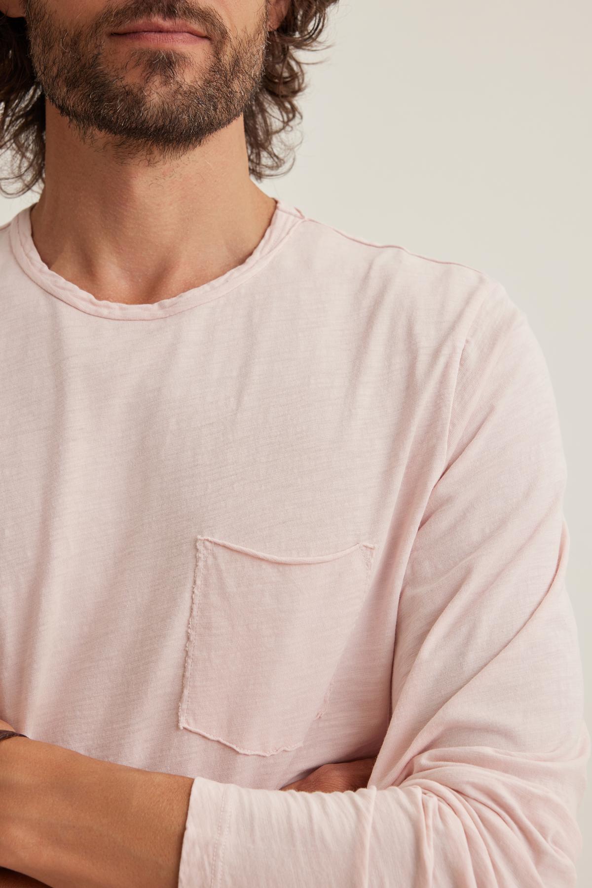 A man confidently stands with crossed arms, wearing Velvet by Graham & Spencer's SIMEON TEE, a light pink long-sleeve shirt made from soft Peruvian cotton, complete with raw-edge details and a chest pocket.-38643350175937