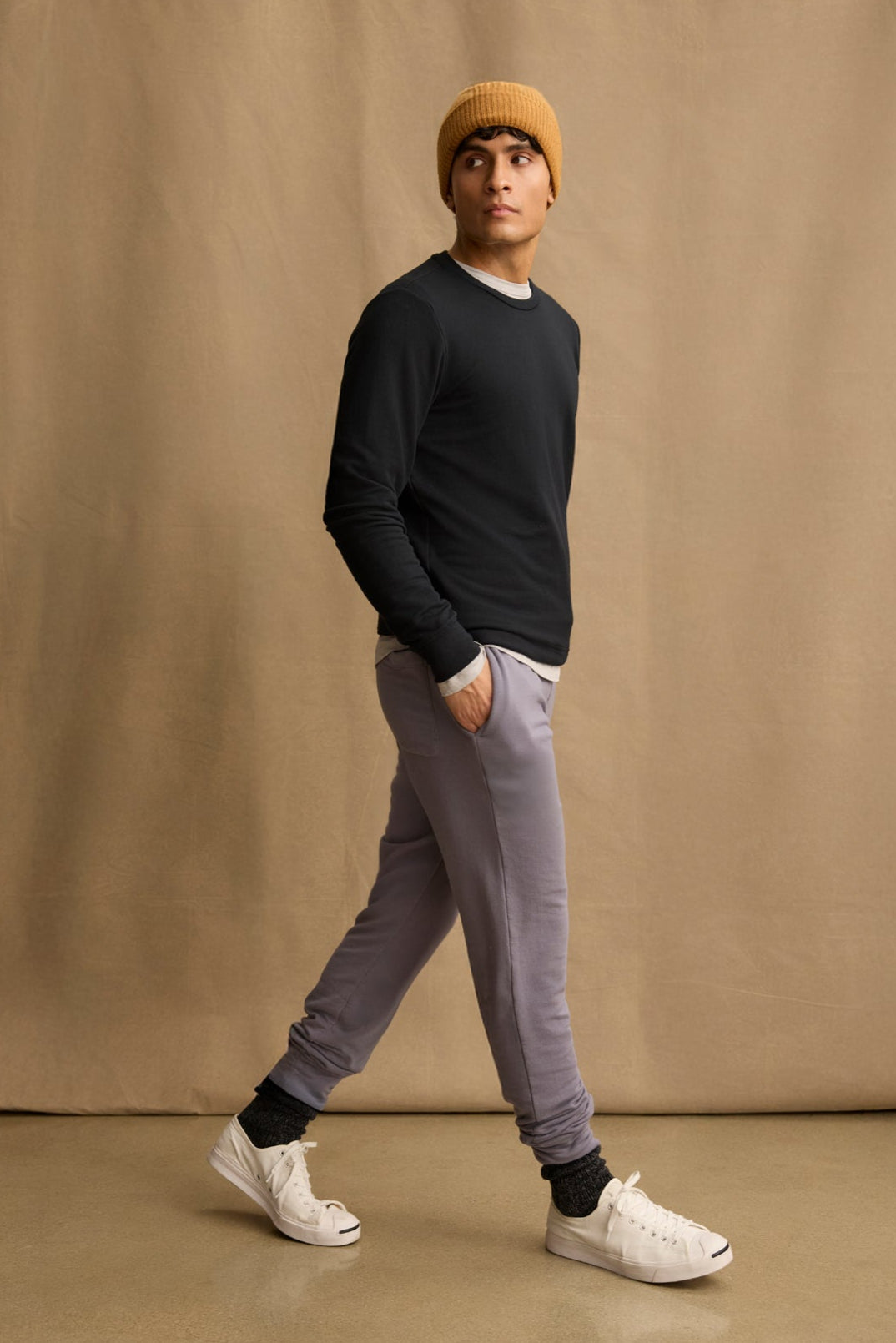 A man in a mustard beanie, black long-sleeve shirt, Velvet by Graham & Spencer CROSBY JOGGER in light gray, dark socks, and white sneakers walks against a beige background.