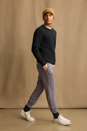 A man in a mustard beanie, black long-sleeve shirt, Velvet by Graham & Spencer CROSBY JOGGER in light gray, dark socks, and white sneakers walks against a beige background.