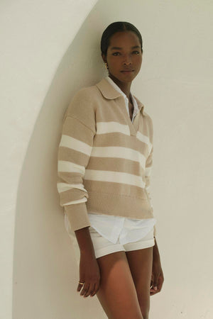 A woman in white shorts and a LUCIE POLO SWEATER by Velvet by Graham & Spencer with stripes leaning against a wall.