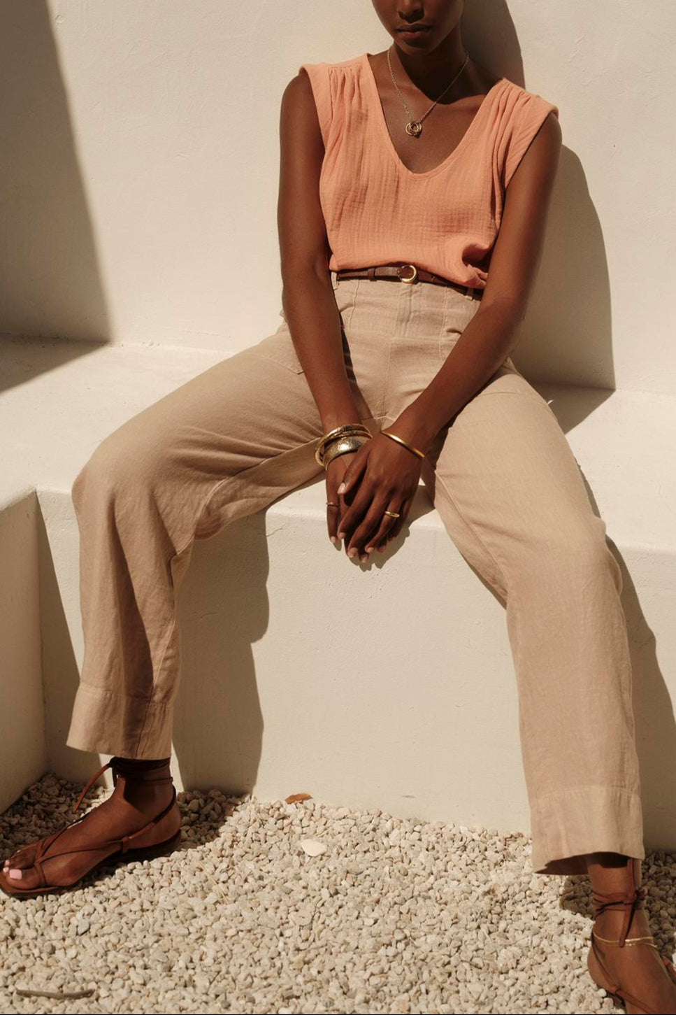 A person wearing the JAYLA COTTON GAUZE TANK TOP by Velvet by Graham & Spencer, along with beige trousers and sandals, sits on a white bench against a white wall. The individual accessorizes with a colorful headband, bracelets, and a necklace.